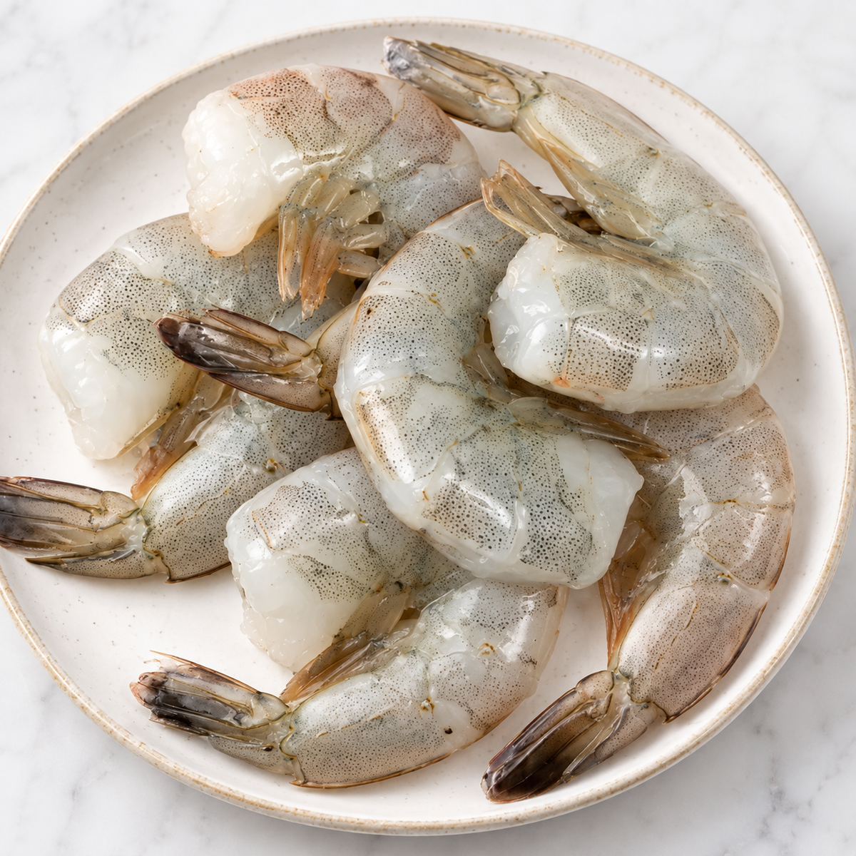 Raw peeled shrimp prawns arranged on a round off-white ceramic plate, photographed from above on a light marble surface.