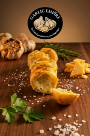 A close-up food scene showing sliced garlic bread rolls on a wooden board with whole garlic, herbs, cheese, coarse salt.